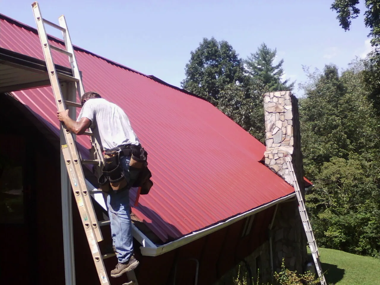 Roofer on a ladder beside a red metal roof and stone chimney — Soffit & Fascia Repair by Roofing Brothers in Ardsley On Hudson